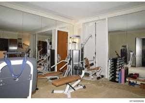This photograph documents a luxury home gym featuring extensive professional-grade fitness equipment including weight machines, free weights, exercise benches, and mirrors. The space appears to be part of a residential property under investigation, w
