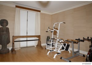 This photograph shows a private home gym or fitness room, featuring multiple exercise machines including a leg press apparatus, pull-up/dip station, and stationary bike. The room has light wood-textured walls, wooden flooring, large windows with whit