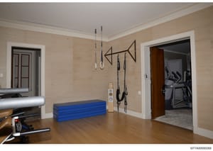 This is a photograph of a private home gymnasium featuring fitness equipment and wall-mounted training apparatus. The room contains suspended training rings, a pull-up bar arrangement, blue exercise mats, cardio equipment, and access to an adjacent f