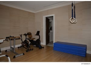 This is an evidence photograph showing a private residential gymnasium containing various exercise equipment including stationary bikes, weight benches, gymnastics mats, and suspended rings. The photo documents the interior of what appears to be a hi