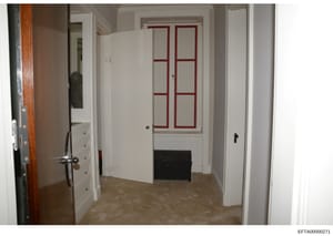 This photograph shows an interior hallway of what appears to be a residential property, featuring white walls, cream-colored flooring, built-in white cabinetry on the left side, and distinctive red-trimmed French doors. The image serves as documentar