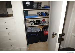 This is an evidence photograph showing the interior of a walk-in closet or storage space containing shelved items, clothing, shoes, bags, and miscellaneous personal belongings. The photo documents the contents of a residential storage area, likely ta