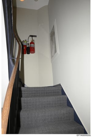 This is a photograph of an interior stairwell in what appears to be a residential or commercial building. The image shows gray carpeted stairs, wooden handrails, white walls, and fire safety equipment mounted on the wall including fire extinguishers 