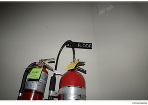 This is a photograph of fire safety equipment (fire extinguishers) installed on a second floor of a building, with location signage and inspection tags visible. The image documents the placement and condition of fire safety apparatus, likely taken du