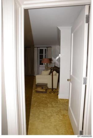 This is a photograph showing an interior hallway and adjacent room in what appears to be a residential property. The image depicts hallway flooring, white trim work, doorways, and a furnished room visible through the open doorway containing a cream-c