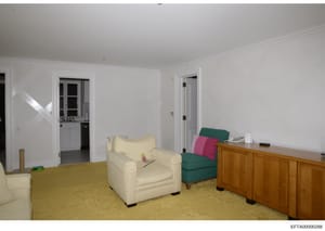 This is a photograph of a residential living room, showing furniture placement and interior design elements. The room features cream-colored walls, a yellow carpet, upholstered seating including a cream armchair and green/pink sectional pieces, woode