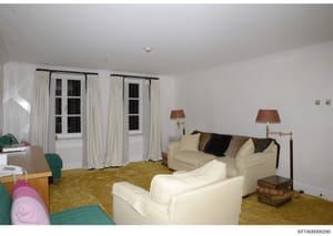 This is a photograph of an interior living room or lounge space, featuring cream-colored upholstered seating, period furnishings, and elegant décor. The room includes French doors with white curtains, wooden furniture, and table lamps with rust-color