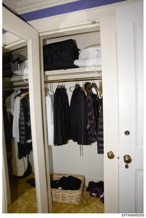 This is a photograph of a residential walk-in closet showing organized clothing storage, including hanging garments, shelved items, and a wicker basket with shoes. The closet features white finishes, brass hardware, and a blue accent stripe on the ce
