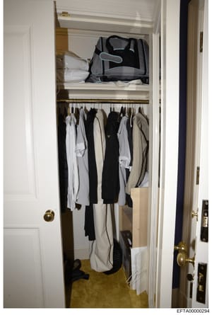This is a photograph of an interior closet space containing hanging clothing and storage items. The closet shows an organized collection of garments in neutral colors (white, black, beige) hanging on a rod, with a black travel bag or duffel on the up