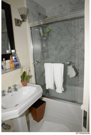 This is a photograph of a modern bathroom interior, showing a white pedestal sink, glass-enclosed shower with gray marble tile, mirror, towel racks, and bathroom amenities. The image appears to be documentation of a residential property, likely from 