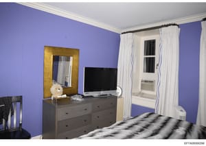 This is a photograph of a bedroom interior featuring distinctive purple-painted walls, a gray dresser with television, a gold-framed mirror, and a bed with patterned bedding. The room includes a window with white curtains, an air conditioning unit, a