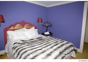 This is a photograph of a bedroom interior, showing a bed with a decorative red headboard, white pillows, and a striped animal-print blanket. The room features purple walls with white crown molding, bedside lamps with red shades, and a Tiffany-style 