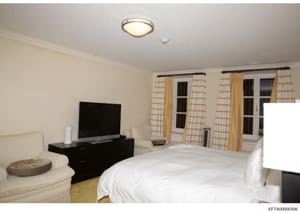This is a photograph of a luxury bedroom interior featuring a king-sized bed, entertainment system, and floor-to-ceiling windows with custom drapes. The room displays high-end furnishings and décor consistent with a wealthy property. The image appear