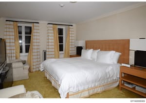 This is a photograph of a luxurious bedroom interior, likely from one of Jeffrey Epstein's properties. The room features a large bed with white linens, tan/gold curtains with geometric patterns, multiple windows, wooden furniture, and contemporary de