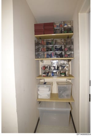 This photograph shows a well-organized storage closet featuring wall-mounted shelving units with multiple clear plastic containers holding various items. The closet contains burgundy storage boxes at the top, several shelves with labeled clear contai