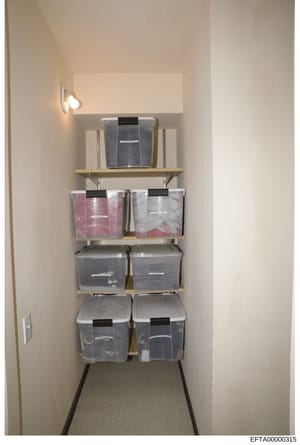 This is an evidence photograph documenting a residential storage closet containing organized plastic storage containers and boxes on wooden shelves. The closet appears to be part of a property search or inventory operation, with various sized clear a