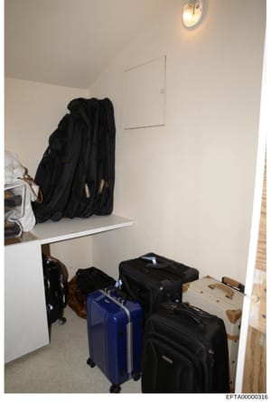 This is a photograph of a residential storage closet containing multiple suitcases, travel bags, and personal clothing items. The image shows various pieces of luggage including a blue rolling suitcase and several black bags, along with bunched black