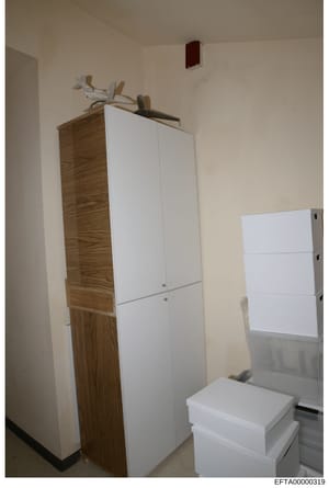 This is an evidence photograph showing the interior of a room containing wooden and white storage cabinets, shelving units, and boxes. The image appears to be from a property search or inventory documentation related to the investigation, showing res