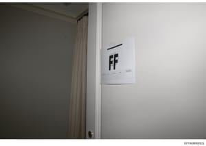 This is an evidence photograph showing the interior of a room with a white document or sign affixed to a white wall. The document displays the letters 'FF' prominently and appears to have a reference grid or measurement scale at the bottom, consisten