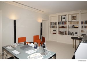 This is a photograph of a contemporary office or residential space featuring a minimalist design with white walls, built-in shelving, and modern furniture. The image shows a work area with a glass desk equipped with computer equipment, orange accent 