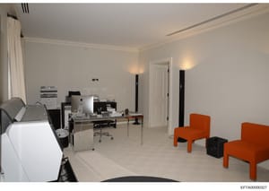 This is a photograph of a contemporary office or residential interior space featuring a work desk with computer equipment, multiple orange upholstered chairs, filing cabinets, and professional lighting. The room appears to be part of an investigation