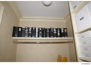 This is an evidence photograph showing a closet or storage area containing approximately 16-18 black binders arranged on a shelf, along with white filing cabinets visible on the right side. The binders appear to be labeled or indexed, suggesting orga