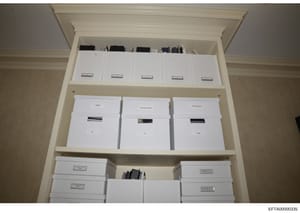 This photograph documents a built-in storage cabinet containing multiple white boxes and file folders, each labeled with identification tags or labels. The boxes appear to be organized evidence storage, likely part of law enforcement documentation an