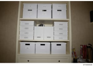 This is an evidence photograph showing a built-in shelving unit containing multiple white storage boxes with various labels and redactions. The boxes appear to be organized evidence or documents related to the investigation, with some labels visible 