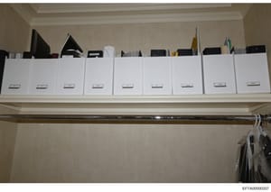 This is an evidence photograph showing a built-in closet shelf containing approximately nine white storage boxes with labels, along with various electronic devices and household items. The boxes appear to be organized and labeled, suggesting this may