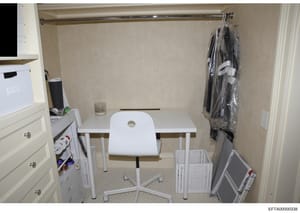 This is a photograph of an interior room, likely a bedroom or office space, showing a simple white desk with a matching white chair, cream-colored walls, a closet area with hanging clothing in plastic, and storage furniture including a dresser with d