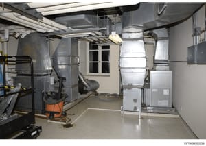 This is a photograph of a basement mechanical/utility room showing HVAC systems, ductwork, piping, and related building infrastructure. The image documents the mechanical systems and basement conditions of a property under investigation, likely taken