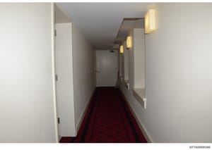 This photograph shows a long, narrow hallway in what appears to be one of Jeffrey Epstein's residences. The hallway features white walls, recessed lighting, multiple closed doors on both sides, and a burgundy patterned carpet. The clean, minimalist d