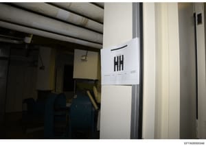 This photograph documents an interior space, likely a basement or utility room, with industrial piping overhead and evidence collection markers visible. A white sheet with tally marks (indicating a counting or tracking system) is affixed to the wall,