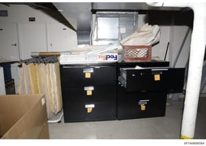 Evidence photograph documenting a storage area or basement space containing two black filing cabinets with yellow evidence tags, assorted boxed materials, window coverings on a rod, and other items. The photograph appears to be from a property search