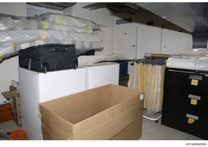 This photograph documents a storage or evidence facility containing various items including rolled materials in plastic wrapping (marked with colored tape), filing cabinets with labeled drawers, boxes, luggage, and other materials. The organized arra