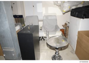 This photograph documents a storage or warehouse room containing various furniture items and boxes, including two upholstered chairs covered in plastic, a black pedestal table, cardboard boxes, plastic-wrapped items, and miscellaneous storage materia