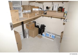 This photograph documents the contents of a storage room or closet space, showing shelving units filled with numerous cardboard boxes, electronic equipment, and miscellaneous items. The image appears to be evidence documentation from the investigatio