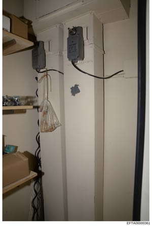 This is a photograph of an interior wall installation showing electrical wiring, two wall-mounted electrical boxes with switches/outlets, a coiled black power cord, and a clear plastic bag hanging from the left electrical box. The image appears to be