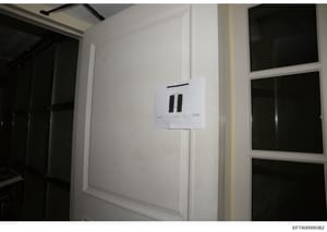 This is an evidence photograph showing an interior room with white walls, dark windows or glass doors, and a white evidence marker board attached to the wall. The image appears to be documentation of a property or room during an investigation, with t