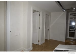 This is a photograph of an interior hallway within a residential or commercial property, showing white painted walls, multiple white doors with brass hardware, wooden flooring, recessed lighting, and what appears to be a mirror or reflective surface 