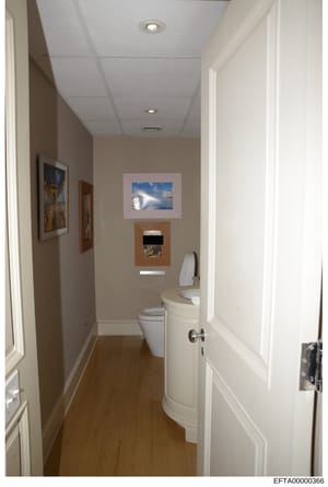 This is a photograph of a narrow, well-maintained bathroom featuring beige and white décor, wood flooring, and multiple framed artworks on the walls. The image appears to be documentation of a property interior, likely taken during an investigation o