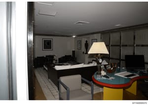 This is a photograph of an interior room showing a modern living or office space with contemporary furnishings, including a desk with a teal-colored top, office chair, lamp, and shelving. The space features minimalist black and white décor with archi