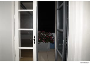 This is a photograph showing an interior view through glass-paneled doors opening to an outdoor patio or balcony area. The image depicts a modern residential or commercial space with white framing, dark-tinted glass panels, and flowering plants visib