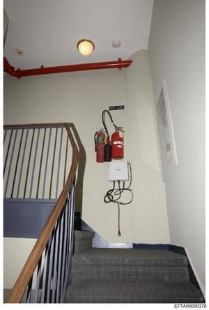 This is an evidence photograph showing an interior stairwell at an Epstein property. The image depicts a wooden-railed staircase with white walls, a red structural beam, recessed lighting, and fire safety equipment mounted on the wall including fire 