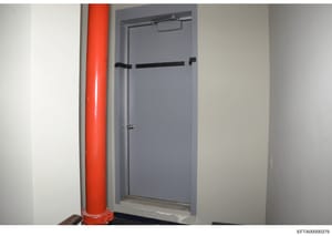 This is a photograph of an interior hallway or corridor showing a gray doorway with black safety bars or restraints, alongside a prominent red structural support column. The image appears to be evidence documentation from a property search or seizure