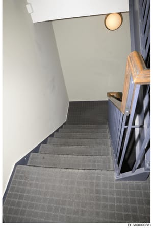 This is a photograph of an interior staircase taken from an elevated angle looking down the stairs. The image shows a functional stairwell with gray carpeted steps, white walls, a wooden handrail on the right side, recessed lighting, and dark blue cu