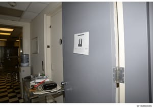 This evidence photograph shows an interior room with light blue/gray walls, fluorescent ceiling, and a metal utility table. A white paper with initials 'JJ' is prominently displayed on the wall, suggesting this may be an office, residence, or facilit