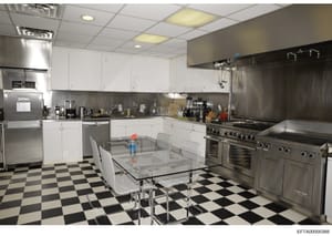 A photograph of a modern, professionally equipped kitchen featuring commercial-grade stainless steel appliances, white cabinetry, and a distinctive black-and-white checkered floor. The space includes a glass-topped dining table, multiple refrigerator