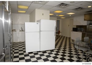 This is a photograph of a large institutional or commercial kitchen space, featuring white cabinetry, two prominent white refrigerators, a black-and-white checkered tile floor, recessed lighting, and basic furnishings. The image appears to be documen