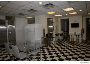 This is an evidence photograph showing the interior of what appears to be a commercial kitchen or employee break room with modern appliances, dining area, and institutional furnishings. The space features stainless steel refrigerators, white cabinetr
